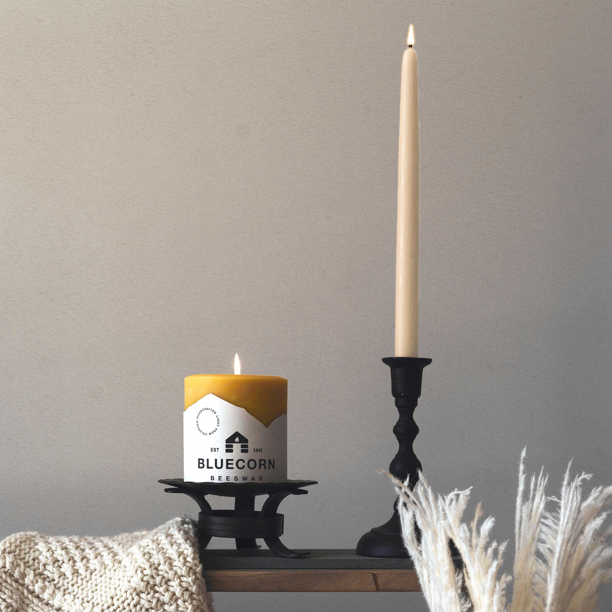Ivory beeswax taper candles next to a yellow candle in a decorative holder on a neutral background.