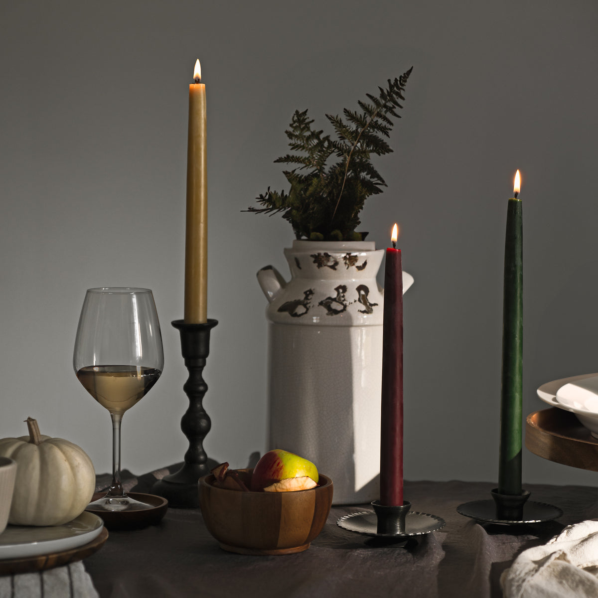 Bluecorn Beeswax hand-dipped burgundy, raw and moss taper candles in Hand Forged taper holder on thanksgiving tablescape with milk jug filled with greenery and glass of white wine.