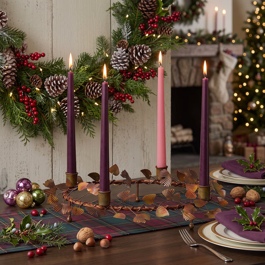 A festive holiday table setting with four taper candles in a copper leaf wreath holder, including two dark purple candles and two light pink candles, styled on a plaid runner with walnuts, red berries, and plates in front of a decorated fireplace.