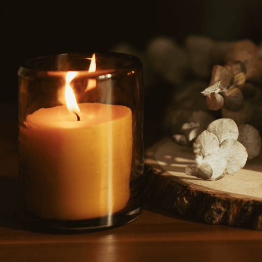 Beeswax jar candle in a blown glass holder with a warm flickering flame, accompanied by dried eucalyptus.