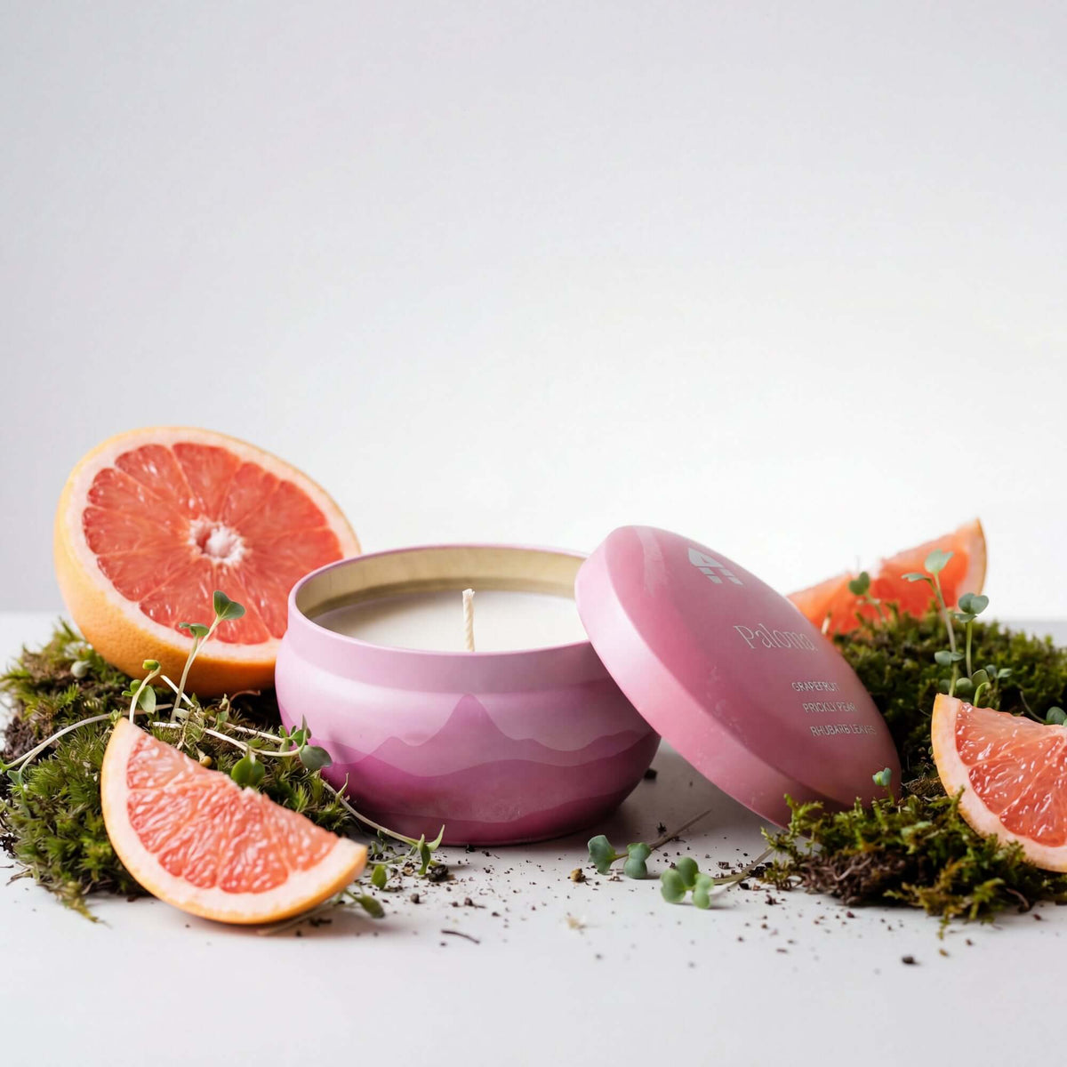 A Bluecorn Aromática 8oz Paloma scented candle tin surrounded by slices of grapefruit and greenery on a white background