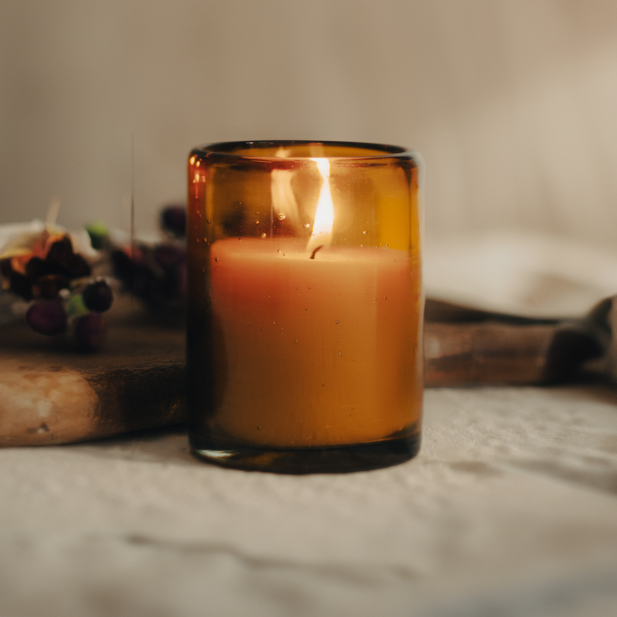 A lit filled candle in an amber glass jar next to a wooden slab with a neutral background