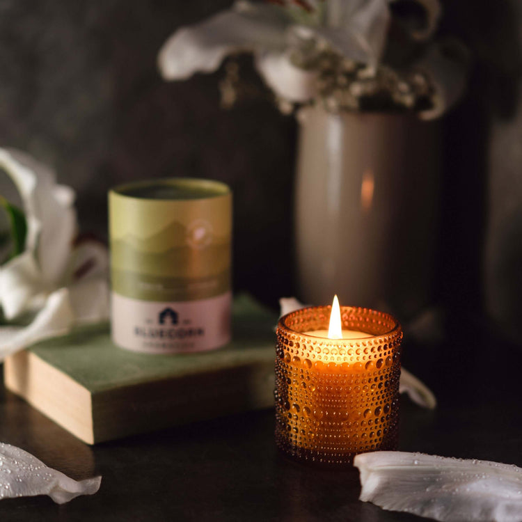 Scented Candles glowing in an amber holder with a decorative candle jar and white lilies background.