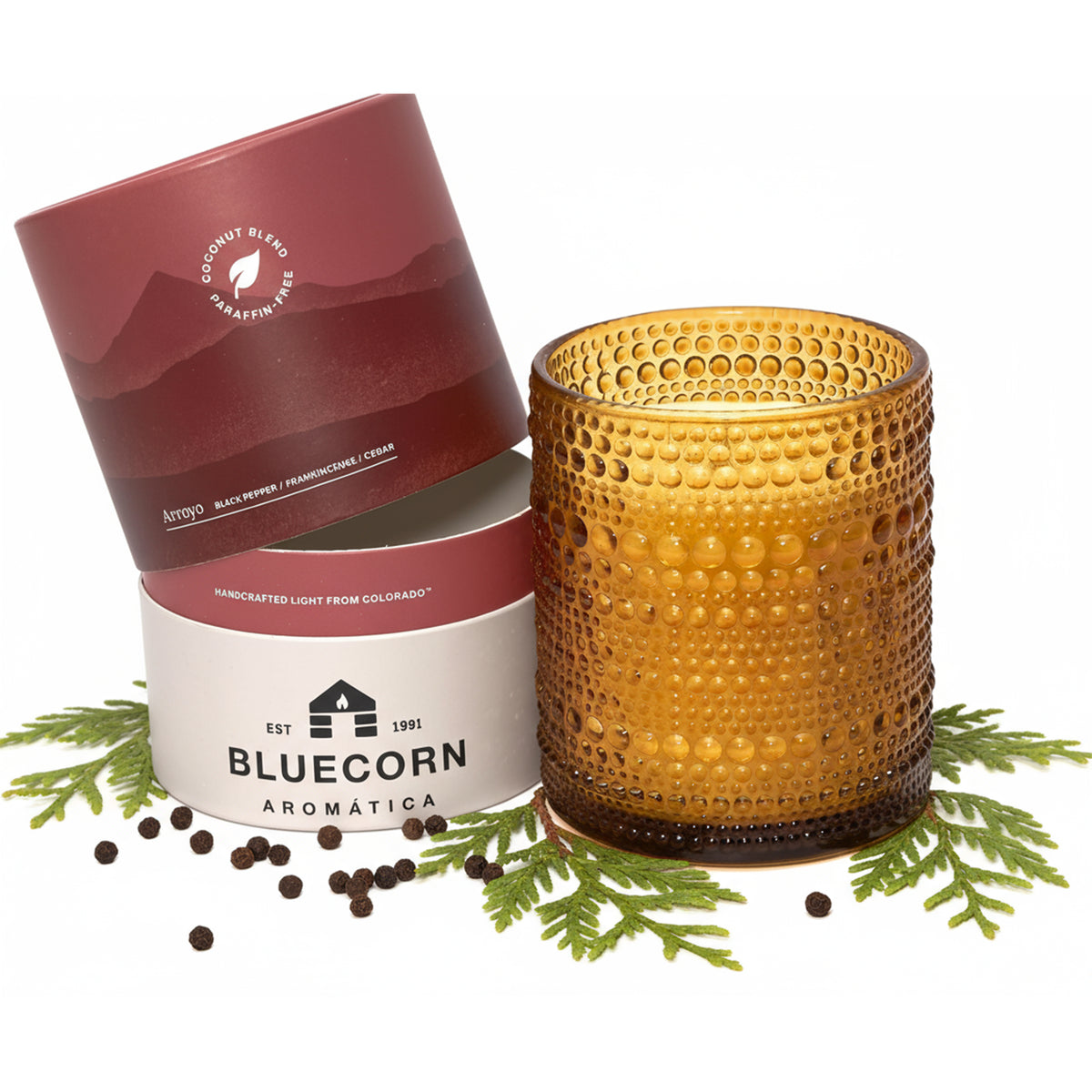 An amber glass candle with a textured dimple finish sits next to its dark red and white cylindrical packaging surrounded by black peppercorns and fresh greenery