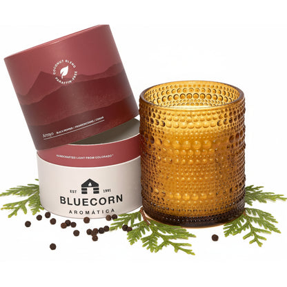 An amber glass candle with a textured dimple finish sits next to its dark red and white cylindrical packaging surrounded by black peppercorns and fresh greenery
