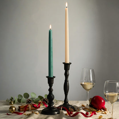 A beeswax taper bundle featuring lit teal and ivory taper candles in black holders, styled with wine glasses, greenery, ribbon and festive holiday décor.