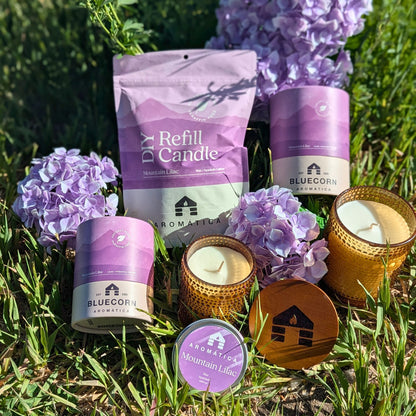 Mountain Lilac scented Bluecorn Aromática Coconut Wax Candles in Sample Tin, Refill Kit, Small & Large glass lying in grass with lilac flowers