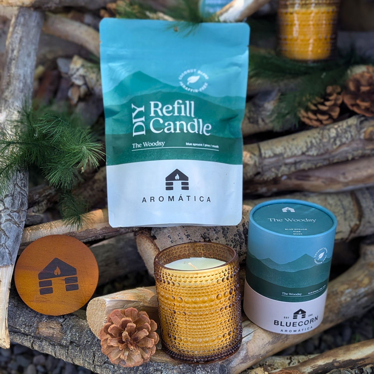 The Woodsy scented Bluecorn Aromática DIY refill kit and filled candle on a wooden background with pinecones