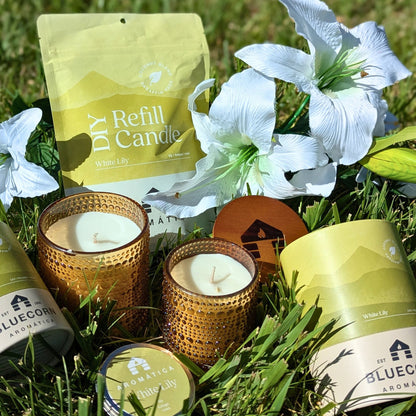 White Lily scented Bluecorn Aromática Coconut Wax Candles in Sample Tin, Refill Kit, Small & Large glass lying in grass with lily flowers