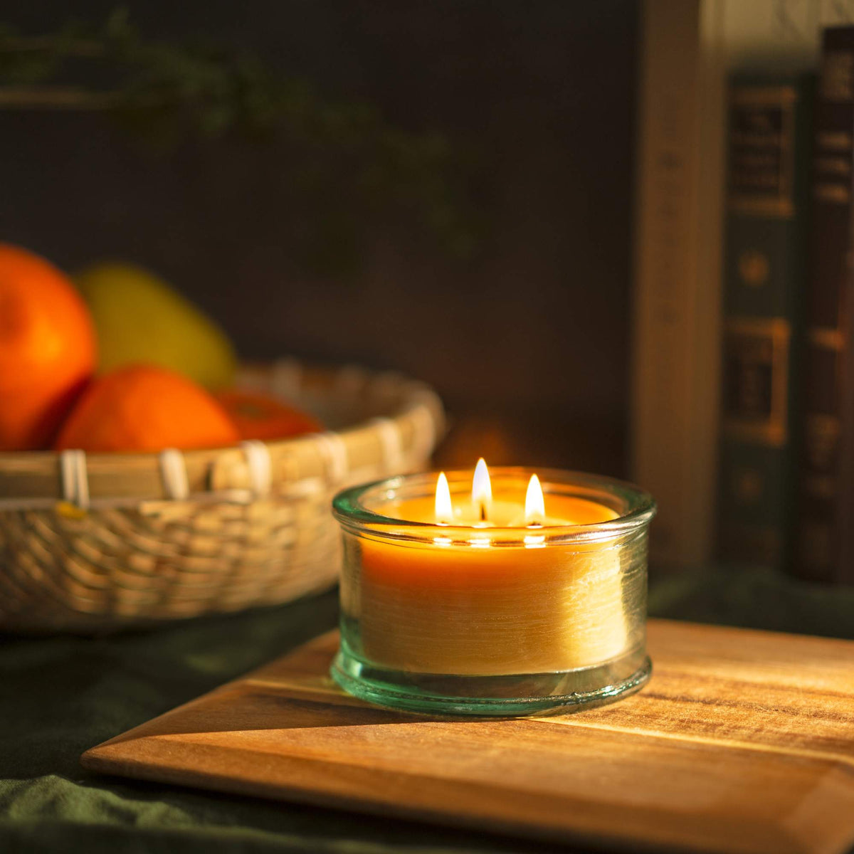 A 100% recycled 3-wick Spanish glass filled candle jar burning on top of a wooden slab with a basket of oranges and books in the background