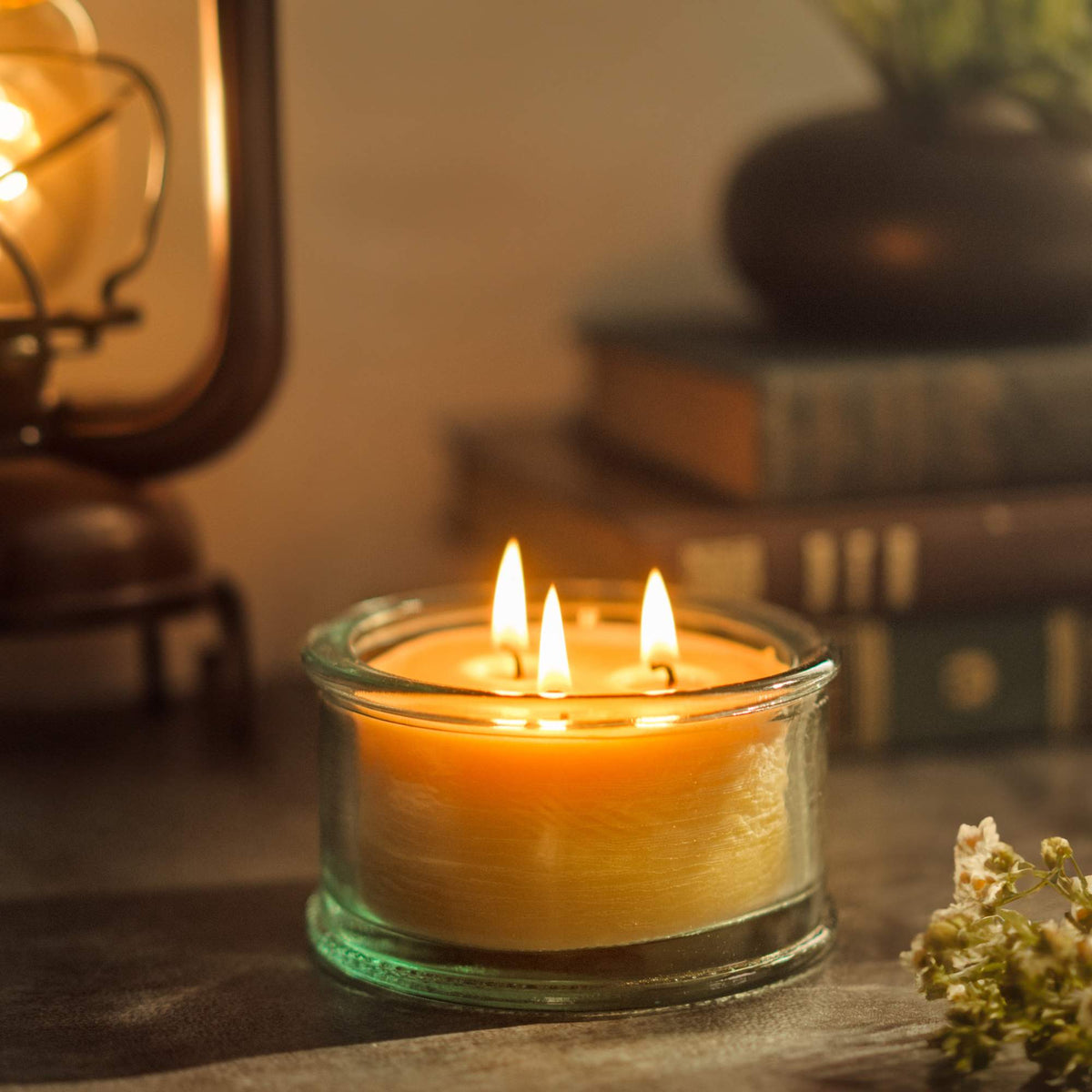 A 100% recycled 3-wick Spanish glass filled candle jar burning on top of a table with dried flowers, a lit lantern and books in the background