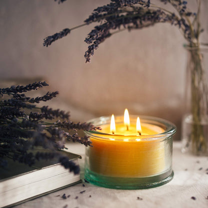 A 100% recycled 3-wick Spanish glass filled candle jar burning on top of a table next to a book and dried lavender