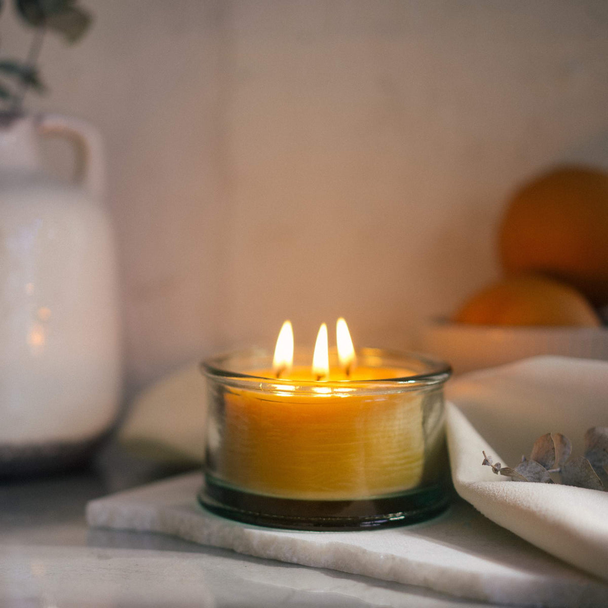 Bluecorn Beeswax scented candle with three wicks glowing in a handmade glass holder, surrounded by oranges and decor.