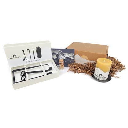 A Pure Beeswax Pillar Candle Gift Set with a gold-foiled gift box, including a 3" x 4" pure beeswax pillar candle, a 4.75" hand-forged pillar base, a candle care toolkit, a refillable apothecary jar of rainbow matches, and a gift card with a handwritten message.