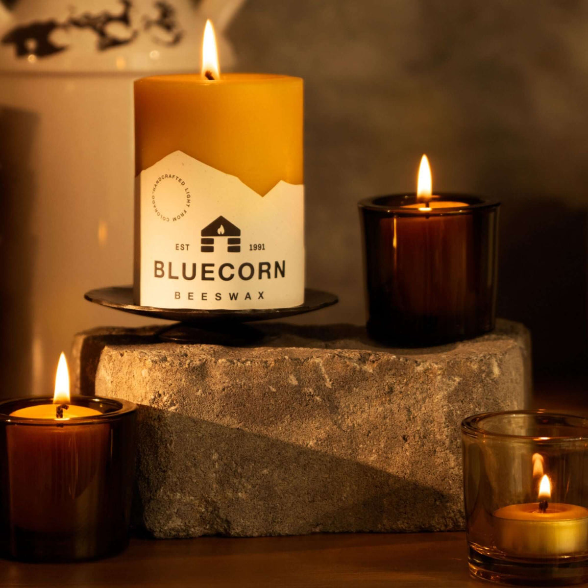 A lit bluecorn beeswax raw pillar candle with a white bluecorn label on top of a stone with lit tea lights and votive candles in votive holders on a wooden surface with a rustic, dark background