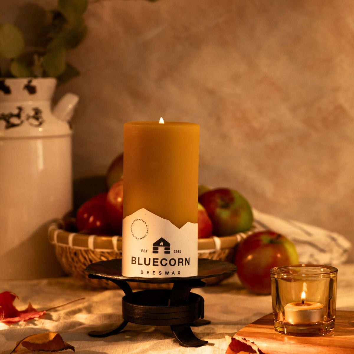 Beeswax pillar candle with Bluecorn label and a lit pure beeswax tea light candle in heavy glass votive holder in a rustic setting with apples and a milk churn.