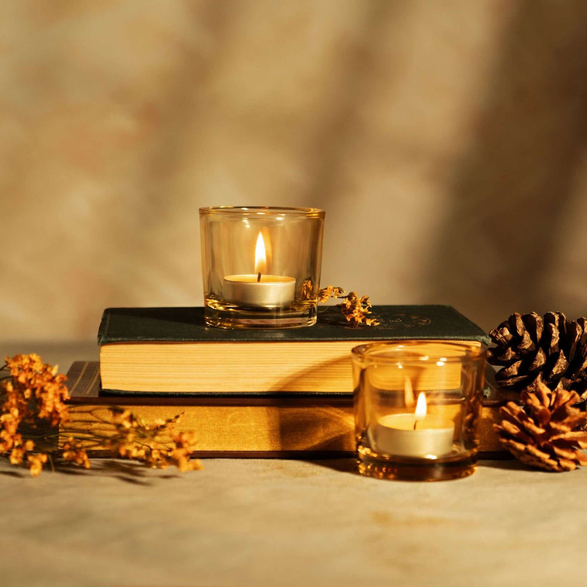 Two lit tea light candles in glass holders on books with a pine cone and dried flowers on a blurred background