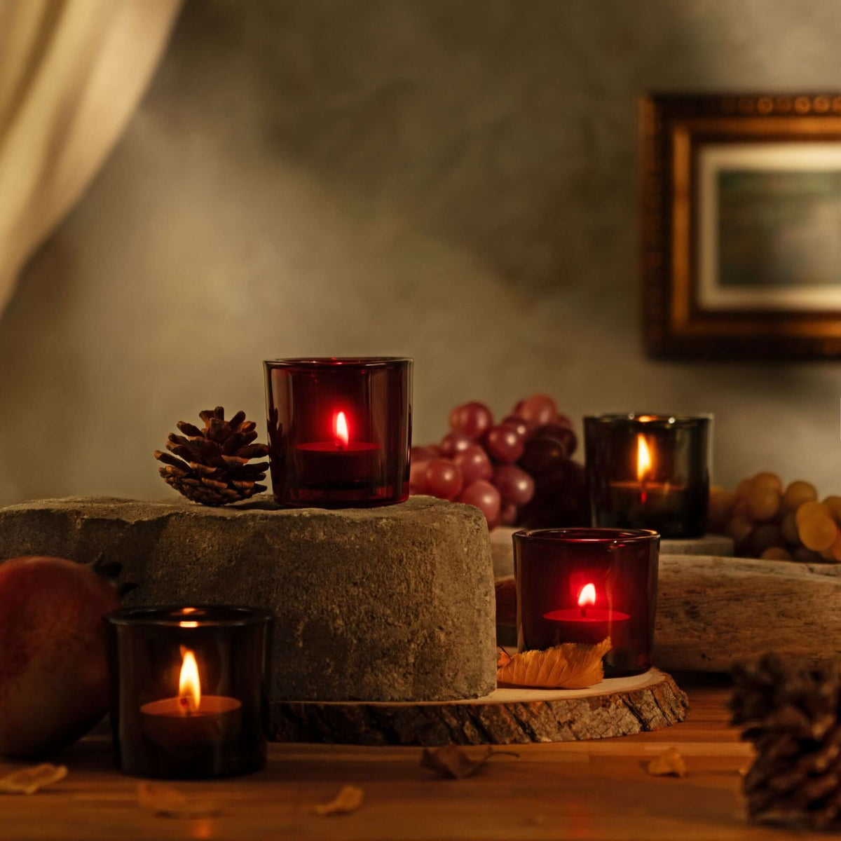 4 lit pure beeswax tea light candles in heavy glass votive holders in decorative setting with pinecones and grapes on a wooden surface