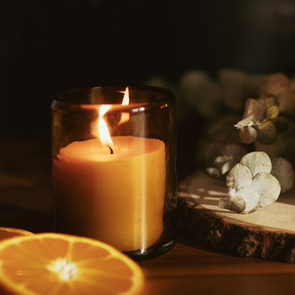 A lit filled candle in an amber glass jar next to a wooden slab with greenery on top and orange slices