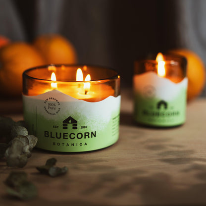 A small and a large 3-wick filled candle in an amber glass jar scented Eucalyptus and Orange. Both candles are lit with green labels stating Bluecorn Botancia on a dark surface with oranges and eucalyptus in the background
