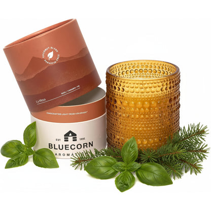 An amber glass candle with a textured dimple finish sits next to its light brown and white cylindrical packaging, styled with fresh basil leaves and pine branches on a white background. 