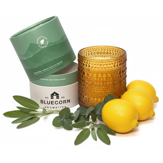 An amber glass candle with a textured dimple finish sits next to its light green and white cylindrical packaging surrounded by fresh greenery and lemons with a white background