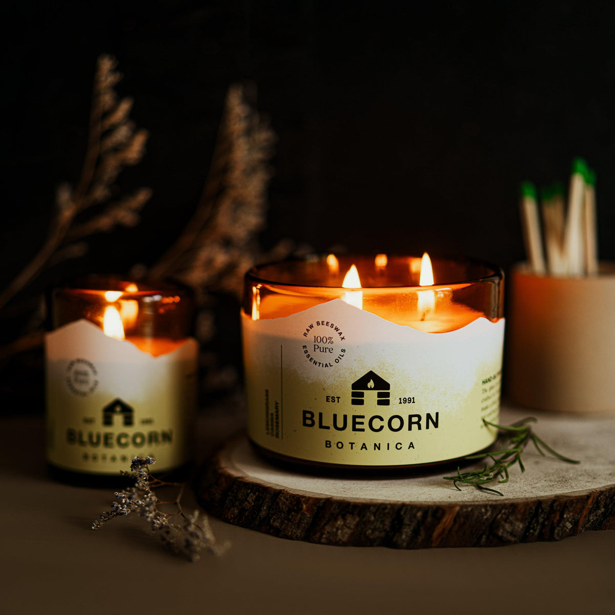 A small and a large 3-wick filled candle in an amber glass jar scented Lemongrass, Cassia and Rosemary. Both candles are lit with yellow labels stating Bluecorn Botancia on a dark surface with matches and dried flowers in the background