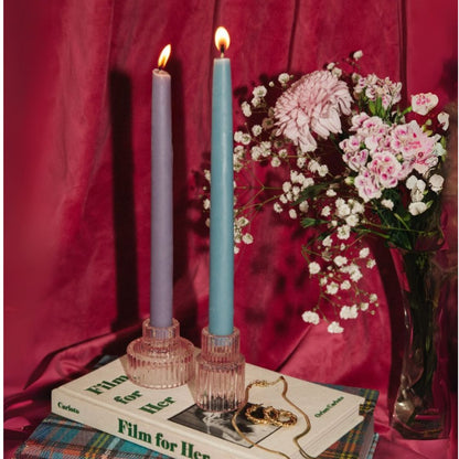 Two lit bluecorn beeswax taper candles in glass holders on books with a bouquet of flowers against a red curtain.