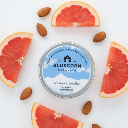 Bluecorn Botanica Beeswax Lotion Bar in Almond Grapefruit. Made with Organic Shea Butter, Organic Cocoa Butter, Avocado Oil, Apricot Oil, Cappings Beeswax, Essential Oils. Using the warmth of your hands, these solids bars will turn into lotions. Can be used all over the body and come in a convenient tin. Features blue Bluecorn Botanica Label on aluminum tin with opened lid showing lotion bar. Lotion bar is cream in color and features Bluecorn cabin.
