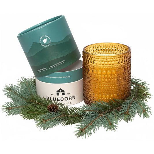 An amber glass candle with a textured dimple finish sits next to its teal and white cylindrical packaging, styled with green pine branches and a pinecone on a white background. 