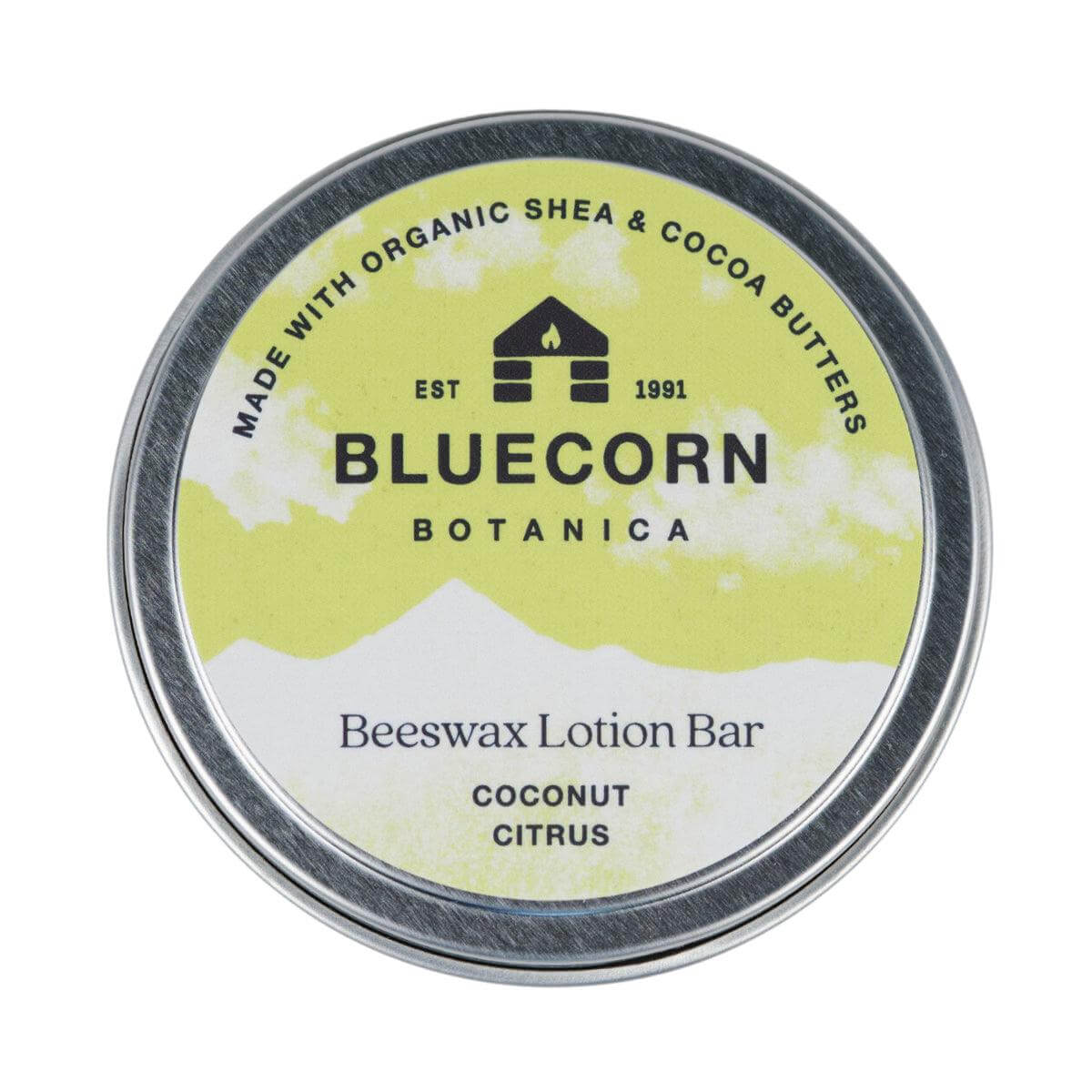 Bluecorn Botanica Beeswax Lotion Bar in Coconut Citrus. Made with Organic Shea Butter, Organic Cocoa Butter, Avocado Oil, Apricot Oil, Cappings Beeswax, Essential Oils. Using the warmth of your hands, these solids bars will turn into lotions. Can be used all over the body and come in a convenient tin. Features green Bluecorn Botanica Label on aluminum tin with opened lid showing lotion bar. Lotion bar is cream in color and features Bluecorn cabin.