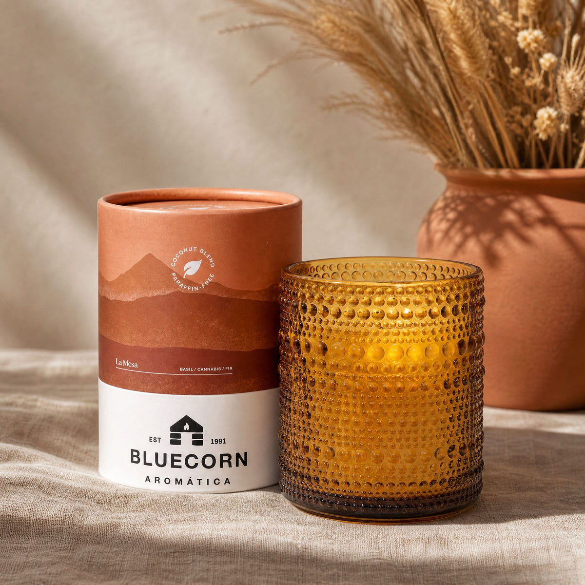 An amber glass candle with a textured dimple finish sits next to its light brown and white cylindrical packaging sitting on top of a textured surface with a terracotta pot filled with dried flowers in the background