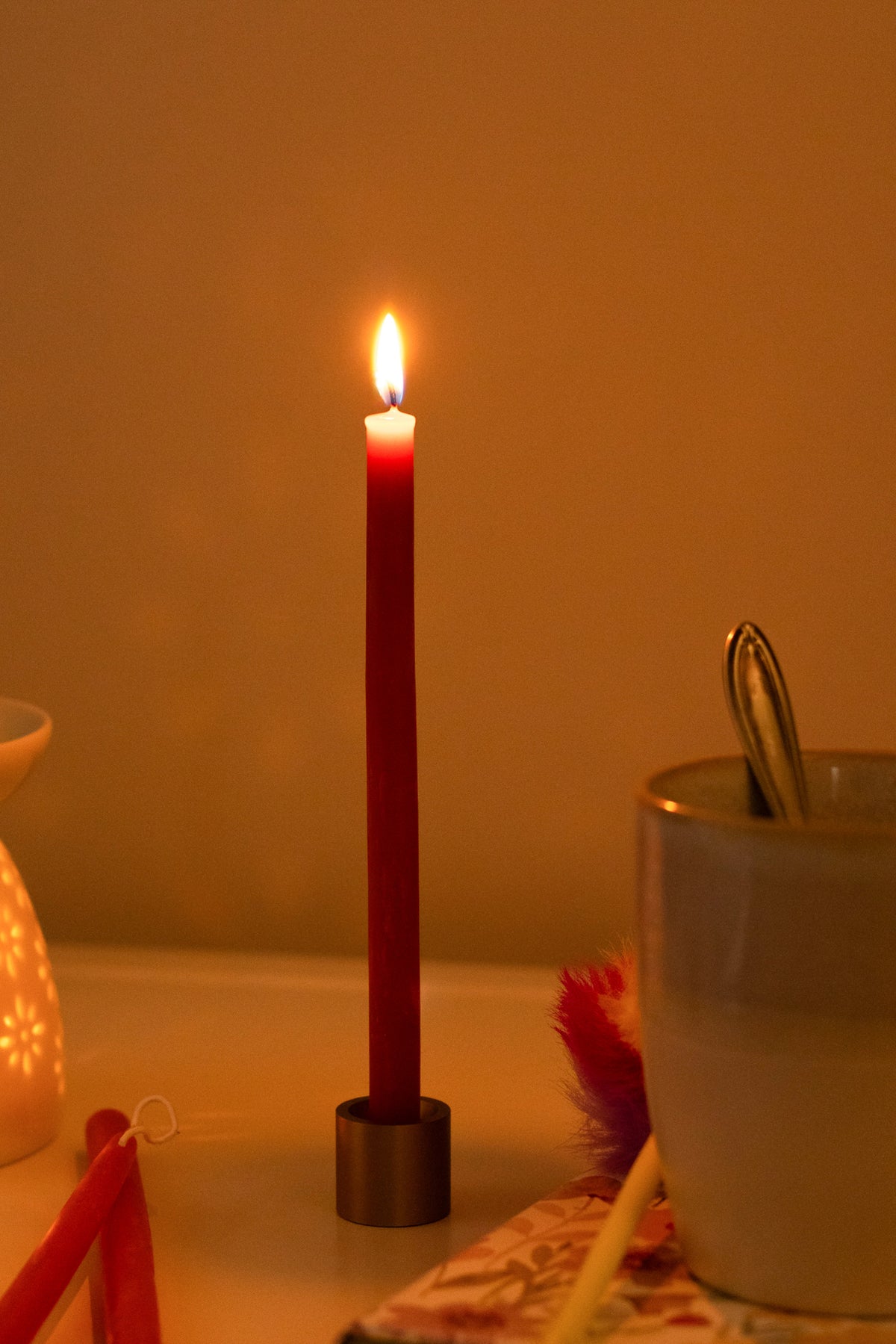 A lit red beeswax hand dipped thin beeswax taper in a brass taper holder on a table next to a white mug and decorative items
