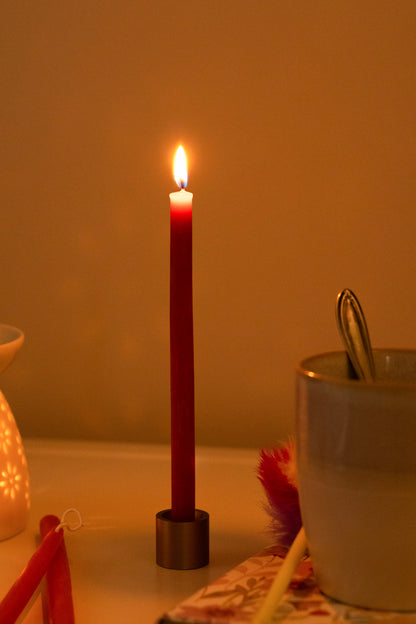 A lit red beeswax hand dipped thin beeswax taper in a brass taper holder on a table next to a white mug and decorative items