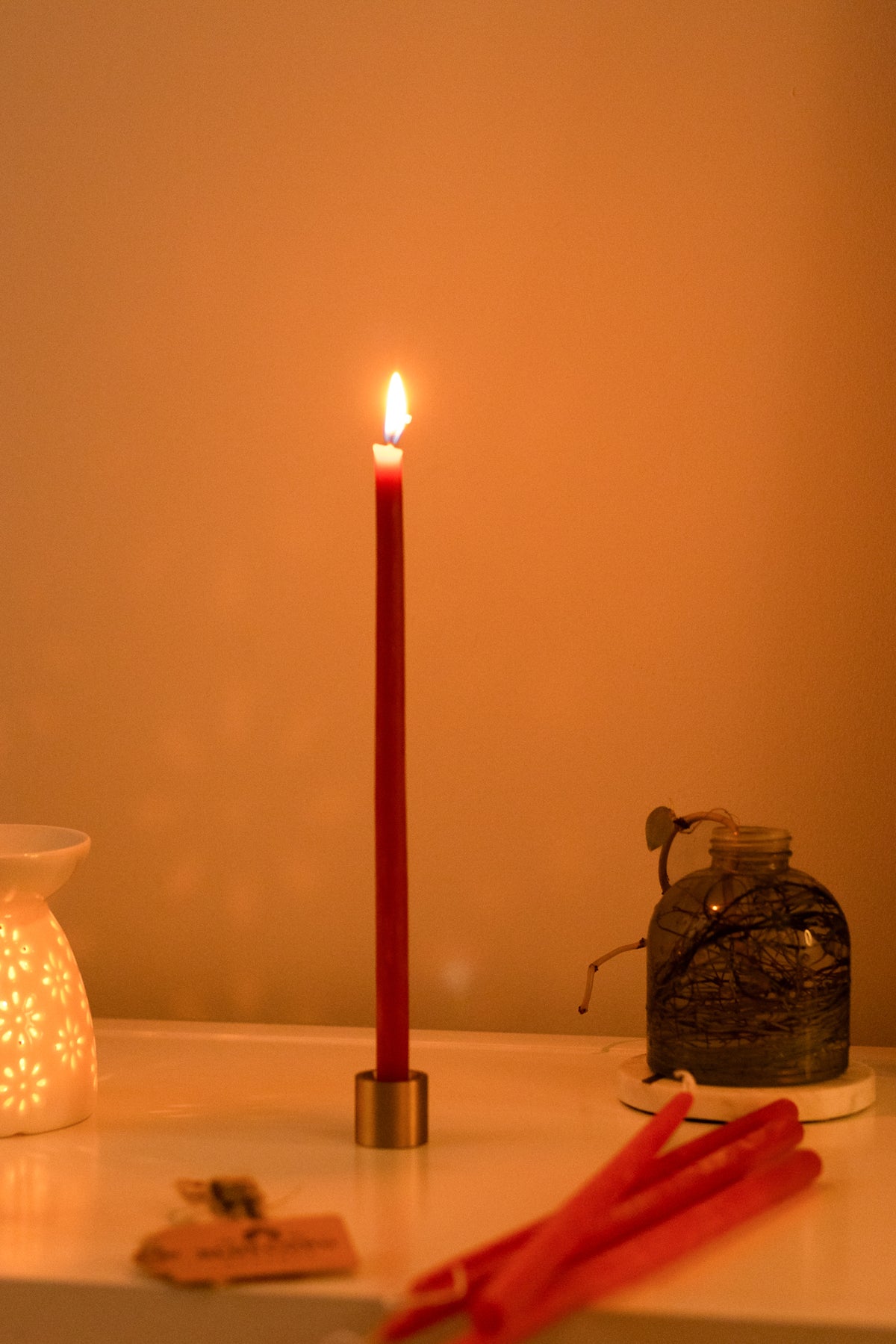 A lit red beeswax hand dipped thin beeswax taper in a brass taper holder on a table next to a white mug and decorative items