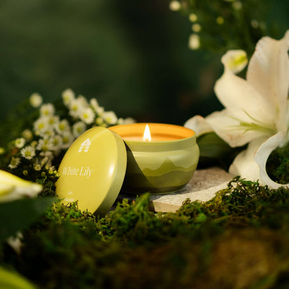 A lit Bluecorn Aromática 8oz White Lily scented candle tin surrounded by Lily flowers and greenery