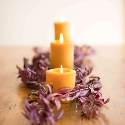 Three lit bluecorn beeswax pillar candles on a decorative wreath with purple leaves on a wooden surface.