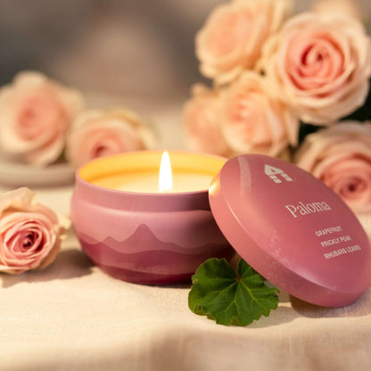 A lit Bluecorn Aromática 8oz Paloma scented candle tin surrounded by pink roses on a soft surface.
