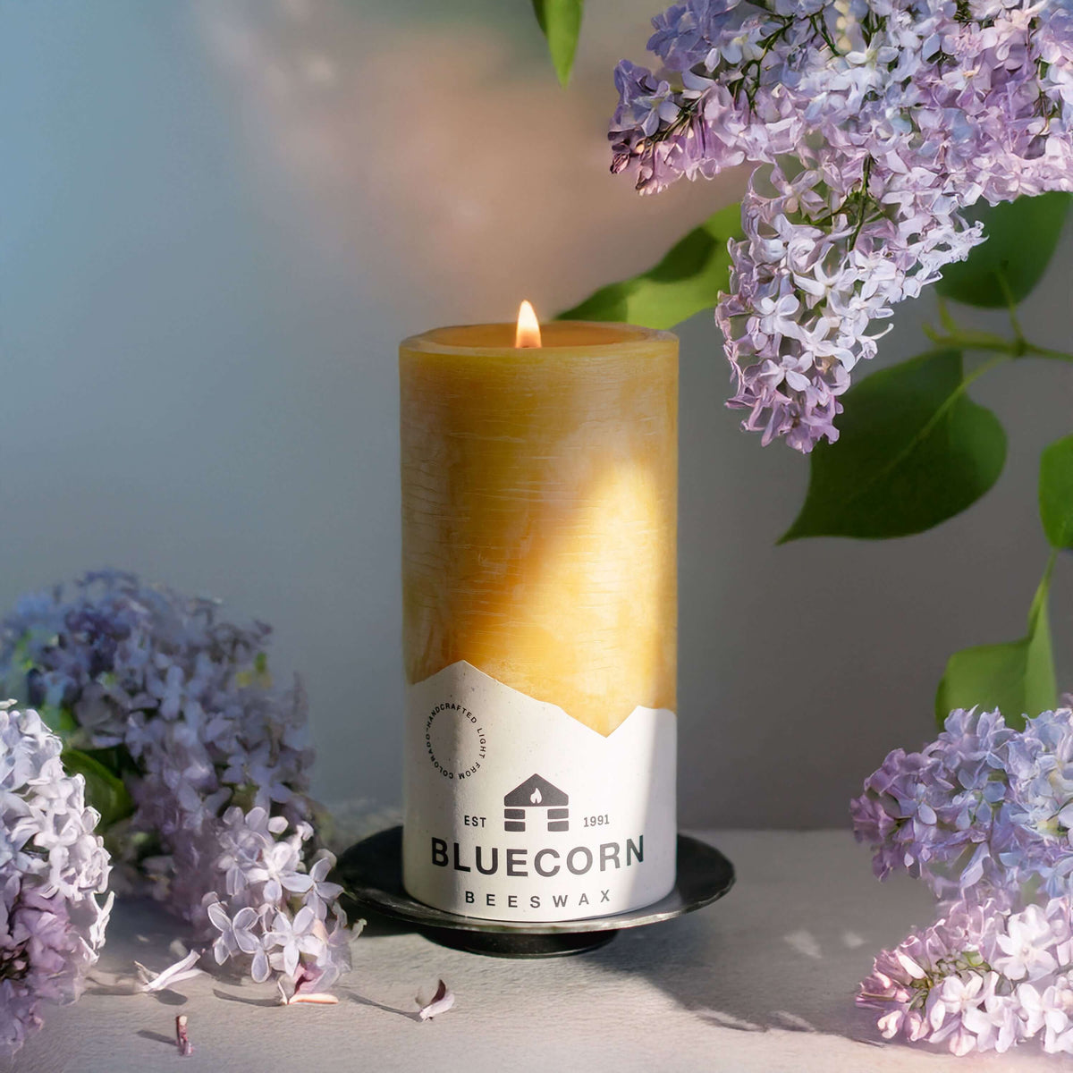 A lit bluecorn beeswax pillar candle with white bluecorn label on top of a forged pillar base surrounded by lilac flowers and a neutral background