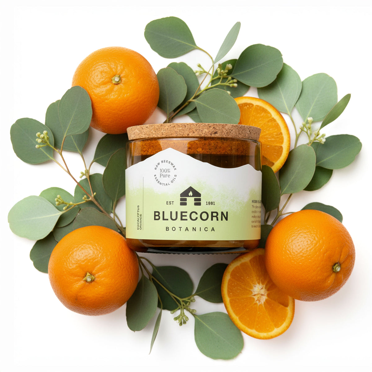A filled jar candle in amber glass centered on a white background, surrounded by whole and sliced oranges and green eucalyptus leaves. The jar has a cork lid and a white label with green and black text.