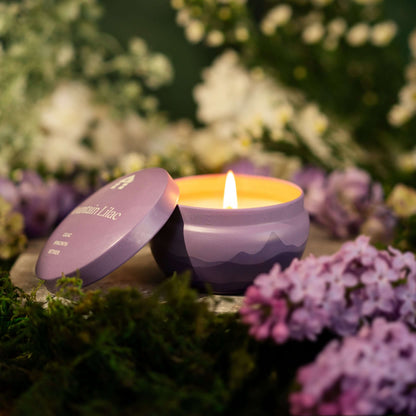 A lit Bluecorn Aromática 8oz Mountain Lilac scented candle tin surrounded by lilac flowers and greenery