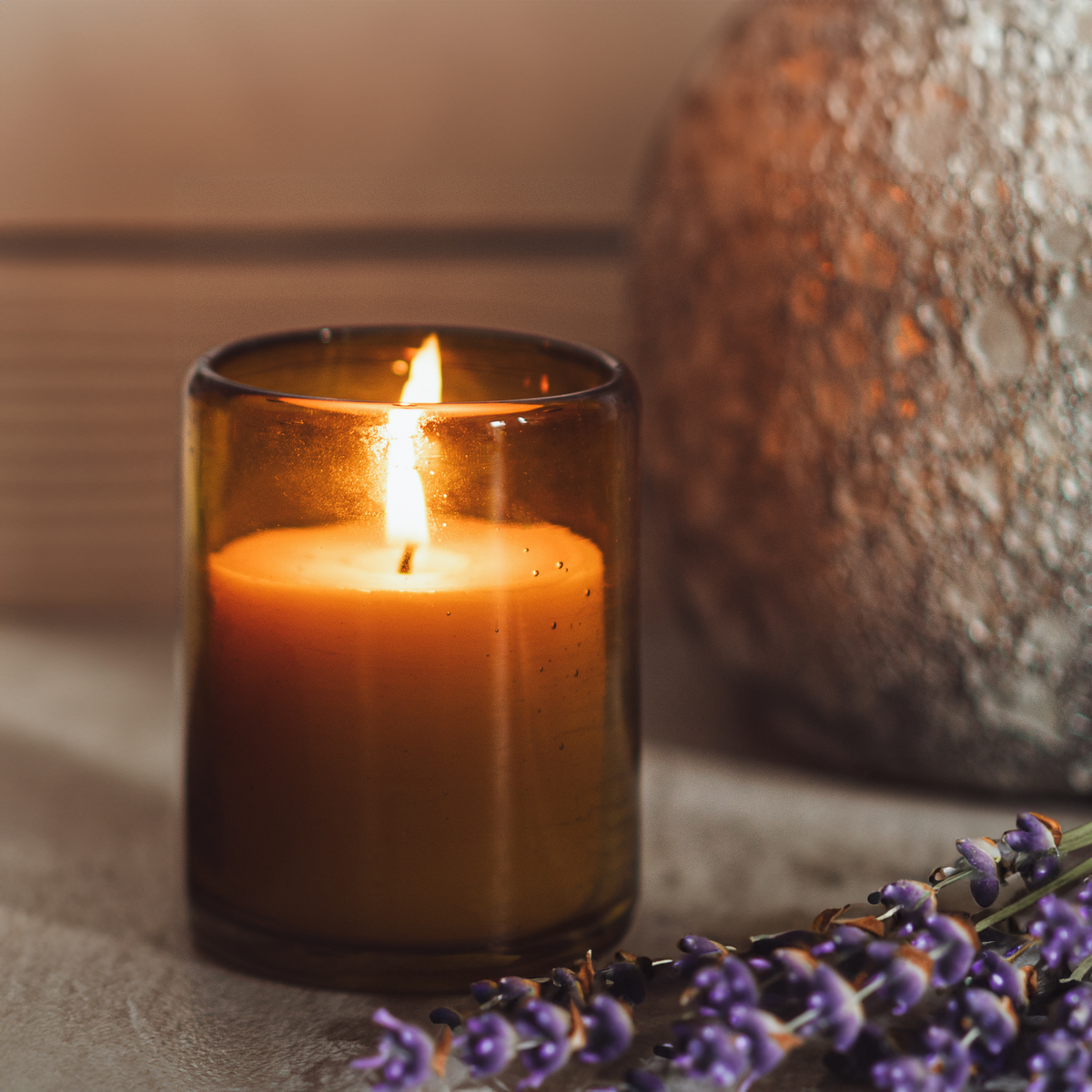 A lit filled candle in an amber glass jar next to dried lavender and a stone vase